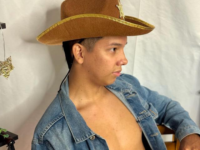 EdwinBlaze, shirtless with a blue denim jacket, wearing a brown cowboy hat, has a brown skin tone, short black hair, and a neutral expression, sitting in a chair.