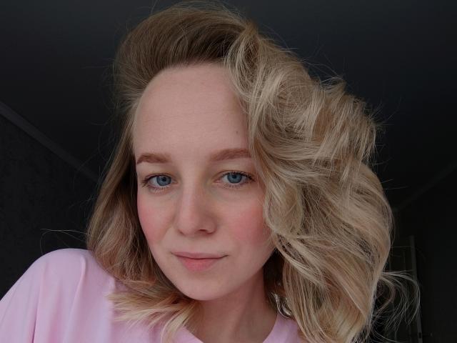 MaryannKellie with blonde, wavy hair, blue eyes, and pink cheeks, wearing a pink shirt, smiling softly. Her tits are not visible, just her face.