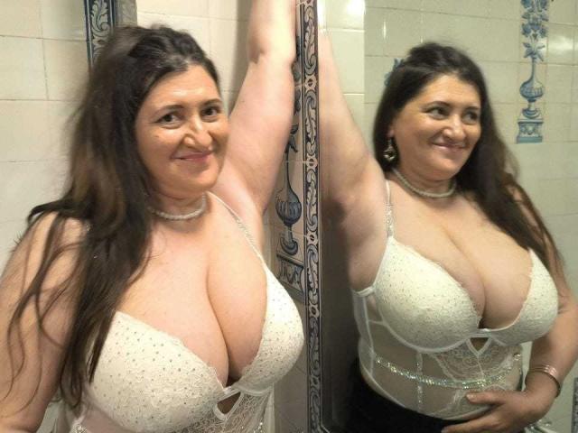 BlissSophia with large tits, wearing a white lace bra, standing against a mirror, smiling, arm raised, long brown hair, choker necklace, reflection showing same.