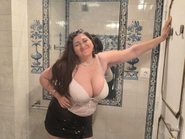 BlissSophia, a curvy woman with big tits, wearing a white lace bra and black skirt, leans against a bathroom wall, smiling, with long brown hair and a pearl necklace.