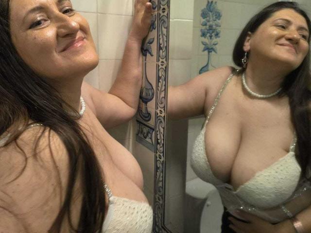 BlissSophia with large tits, wearing a white lace bra and pearl necklace, smiling in a mirror selfie. Her long brown hair, and reflection are visible.