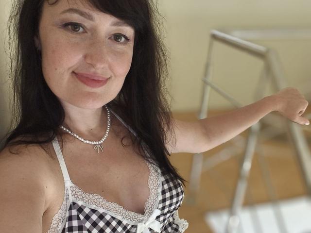 NathalyStars with long black hair, light skin, and freckles, wearing a checkered bra with lace, pearl necklace, smiling, right arm raised, showing small tits and a cute face.