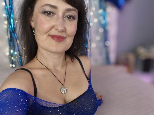 NathalyStars with dark hair, light skin, wearing a blue mesh top that shows her tits, black bra, necklace, smiling, red lipstick, and earrings. Bokeh background.