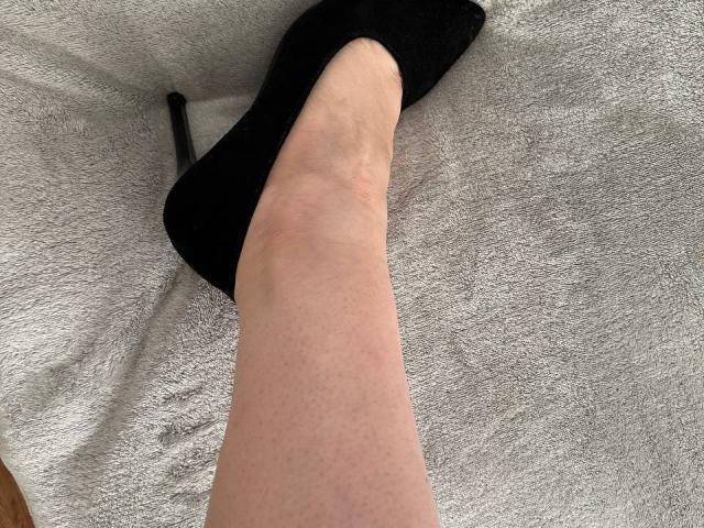 MiraGi's pale, slightly hairy leg with a visible vein, wearing a black, pointed-toe, high-heel shoe on a soft, white, textured surface.