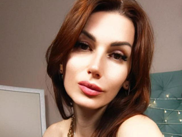 IsabelKiss has brown hair, full lips with pink lipstick, and a slight smirk. She's looking at the camera with a sexy, confident expression. Boobs are partially visible.