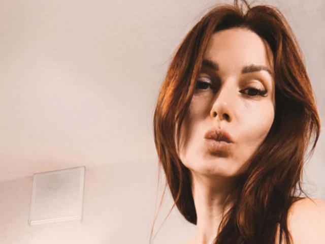 IsabelKiss has brown hair, pouty lips, and subtle makeup. She's looking to the side with a slightly annoyed or bored expression. Her tits are not visible.