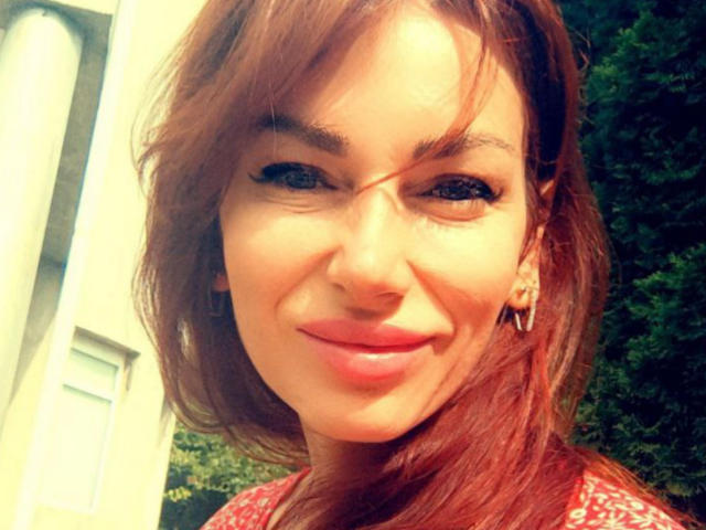 IsabelKiss has red hair, a nose piercing, and a subtle smile with pink lips. She's wearing a red patterned top and has small triangular earrings.