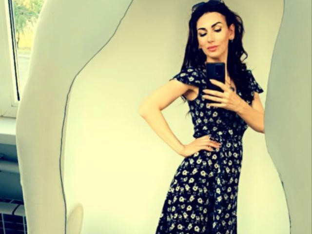 IsabelKiss with dark hair in a floral dress, holding a phone, posing with hand on hip, reflection in a wavy mirror, looking confident, smiling slightly.