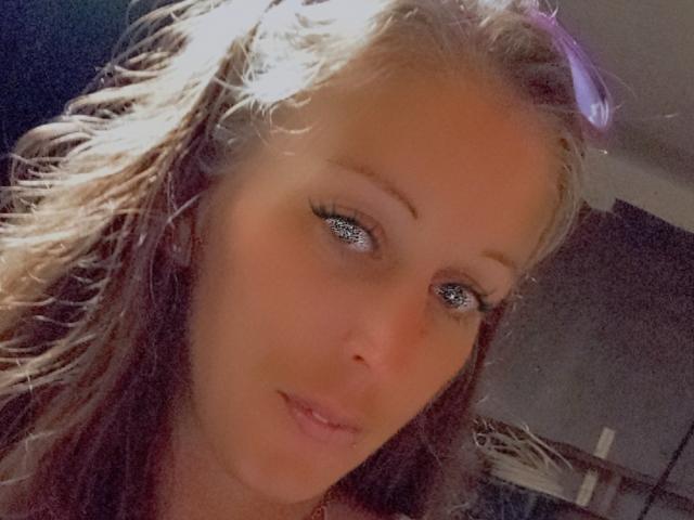 PlayfulRyder has blonde, wavy hair, blue eyes, and a slight smile. Her eyelashes are long and she's wearing purple sunglasses on her head. Her skin is fair.