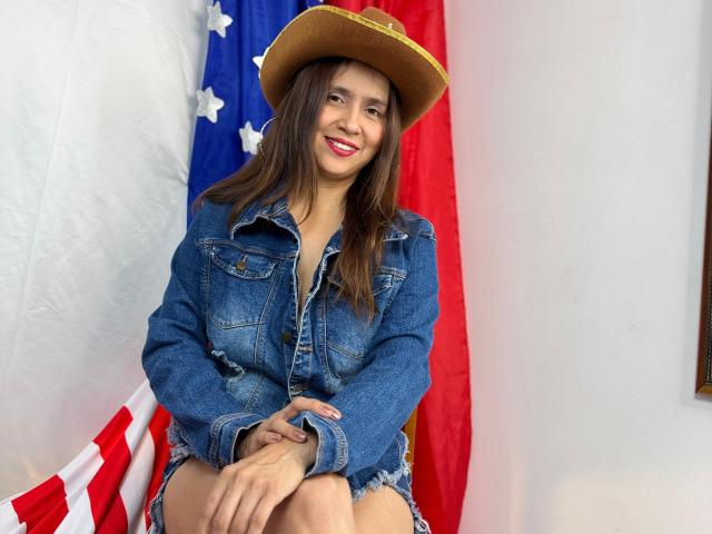 Heidy Wells, con lunghi capelli castani, indossa una giacca di jeans, pantaloncini e un cappello da cowboy, sorride, con le mani giunte, sullo sfondo si vede la bandiera americana, con colori rosso e bianco..