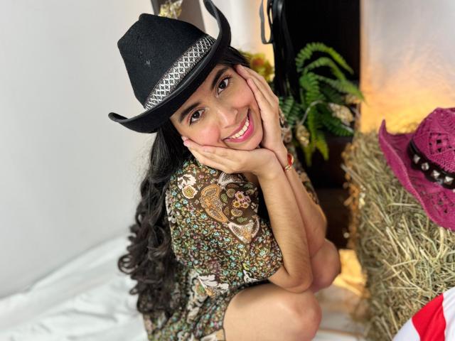 AnnaAlvarez, smiling, wearing a black cowboy hat with silver trim, hands on face, brown floral dress, long black hair, sitting on hay, pink cowboy hat in background.