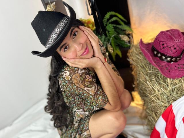 Anna Alvarez mit langem, schwarzem Haar, die einen schwarzen Cowboyhut, ein geblümtes Hemd und einen pinken Cowboyhut trägt, lächelt auf Heu, mit den Händen vor dem Gesicht, braunen Augen und pinken Lippen..