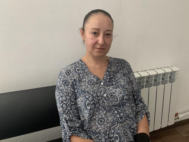 MelissaShell has a medium build, wearing a blue and white paisley shirt, with her brown hair in a tight bun, sitting next to a radiator, smiling slightly.