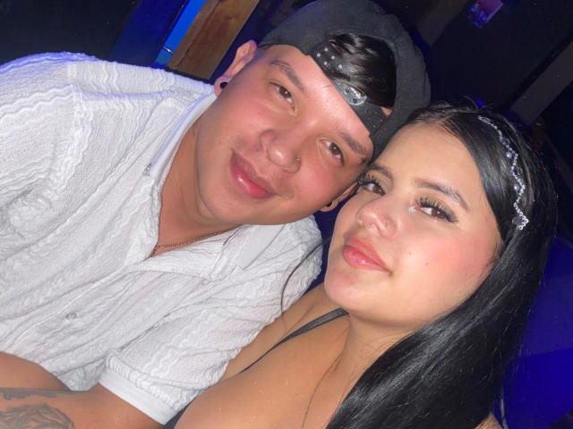SofiaAndLian with black hair, pink lipstick, and large tits, leaning on Lian, who has a white shirt, black cap, and small nose ring, smiling at the camera.