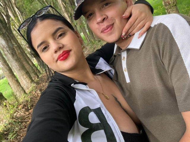 SofiaAndLian with red lipstick, black hair in a ponytail, and a black and white zip-up, showing her tits, arms around Lian in a brown polo, forest background.