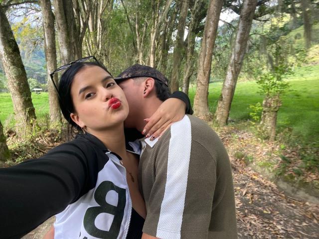 SofiaAndLian with red lipstick, black hair, and a white zip-up jacket, has her arm around Lian, who is nuzzling her neck, wearing a brown and white shirt.