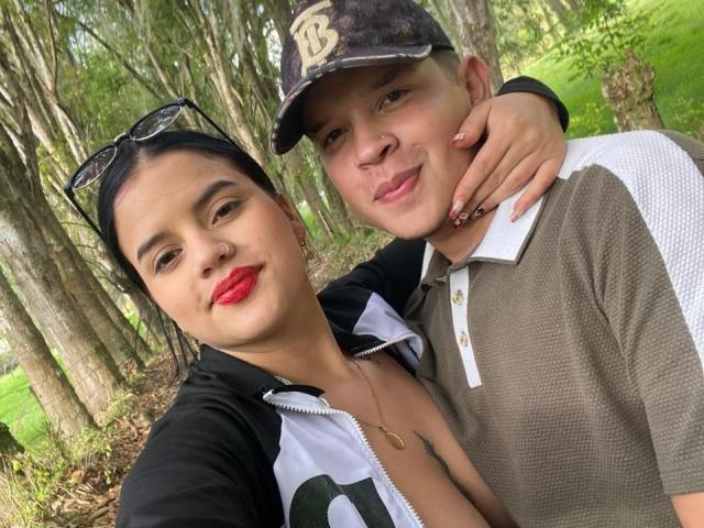SofiaAndLian with red lipstick, black zip-up, and glasses on head, arm around Lian in brown cap, white polo, forest selfie, Lian's nose slightly curved, both smiling.