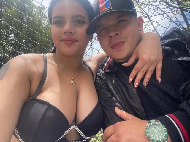 SofiaAndLian with big tits in a black bra, arm around Lian, wearing a cap and black jacket, both smiling, outside, Lian's hand on SofiaAndLian's shoulder, sunny.