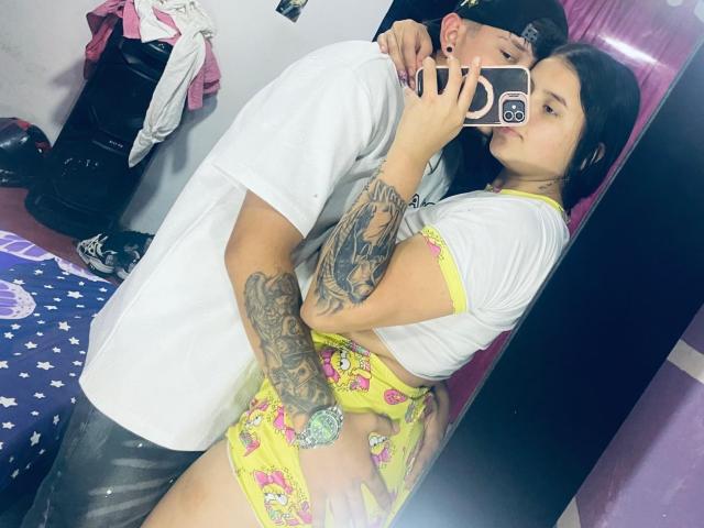 SofiaAndLian, tattooed, wearing yellow shorts, white tee, and cap, being grabbed by Lian's hand on her ass, taking a mirror selfie with Lian's phone.