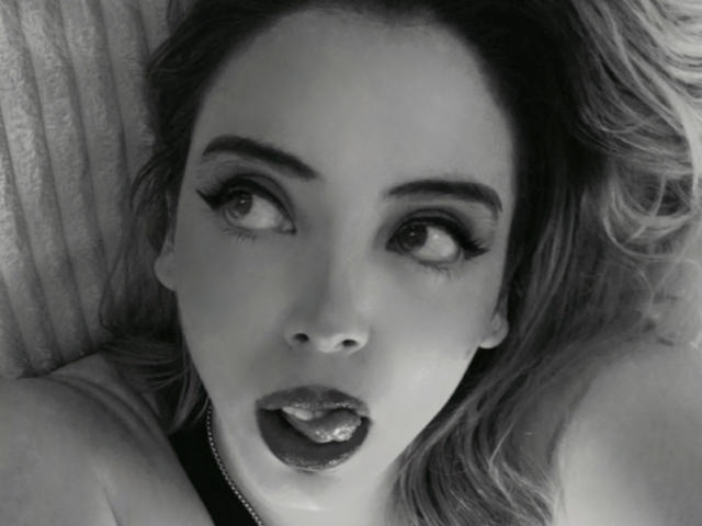Rosalinnnes has her eyes wide open and her tongue slightly out, looking surprised. Her makeup includes thick, long eyelashes. Her mouth is slightly open, showing her upper teeth.