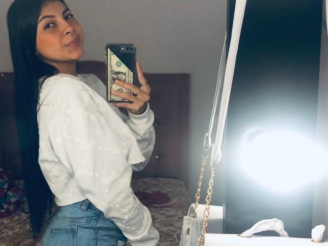 AliceSaadi with long black hair, white sweater, and blue jeans, holding a phone with a dollar bill case, taking a mirror selfie, showing her ass.