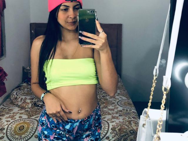 AliceSaadi with long black hair, neon green strapless top, pink hat, blue patterned pants, and belly piercing, taking a mirror selfie in a bedroom.