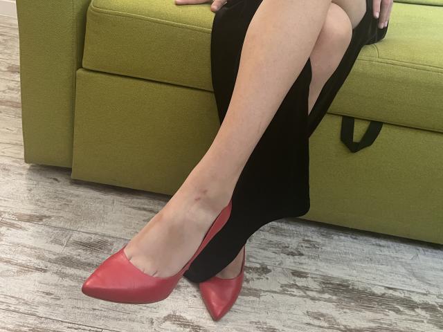 Millinia's legs in a black dress with a cutout, wearing red pointed-toe heels, sitting on a green sofa with wooden floor. She has a small bruise on her left ankle.