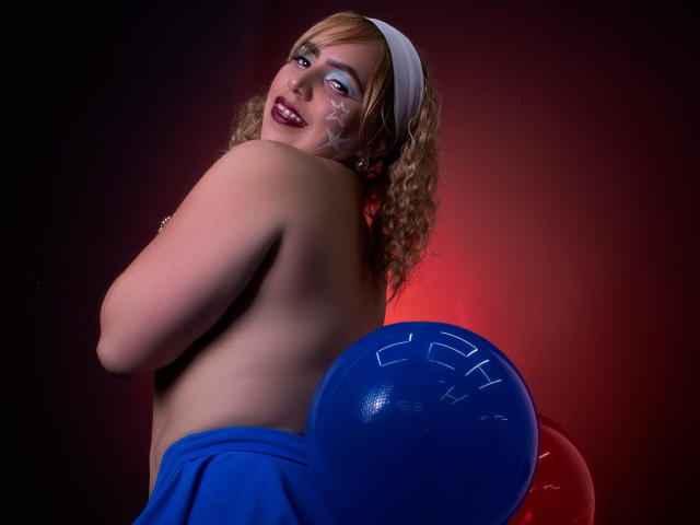 ZaharaBennet, topless with curly blonde hair, white headband, blue makeup, holding blue balloon, smiling, red and black background, visible face and upper body, wearing blue skirt, tits visible.
