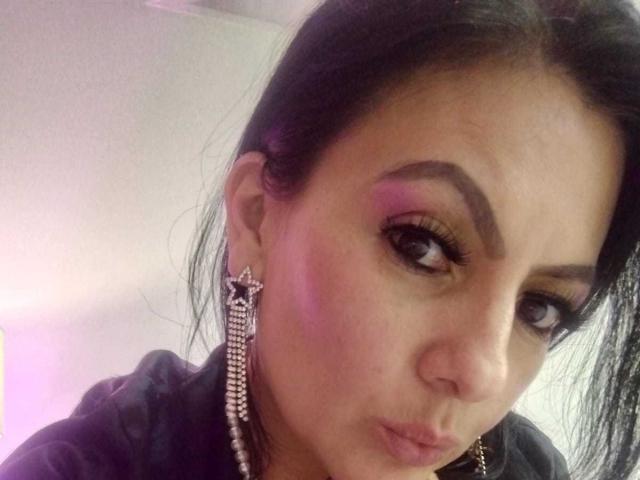 JakyKidman with dark hair, purple eyeshadow, and long eyelashes, wearing star-shaped earrings and a black shirt, pouting her lips. Her tits are small, and her ass is invisible.