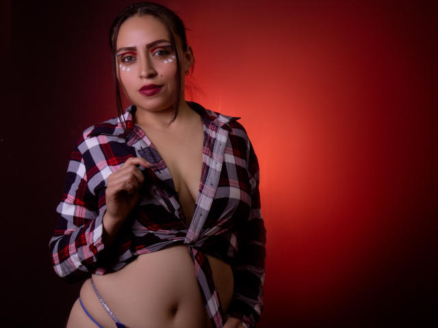 AliceBlackx con una camicia a quadri annodata sotto il seno, che lascia intravedere un po' il sedere, indossa uno slip blu, guarda la fotocamera con un trucco occhi rosso e nero..