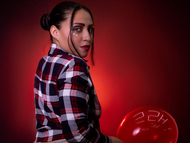 AliceBlackx with dark hair in a bun, wearing a red-black-white plaid shirt, holding a red balloon, looking back with a slight smile, red gradient background.