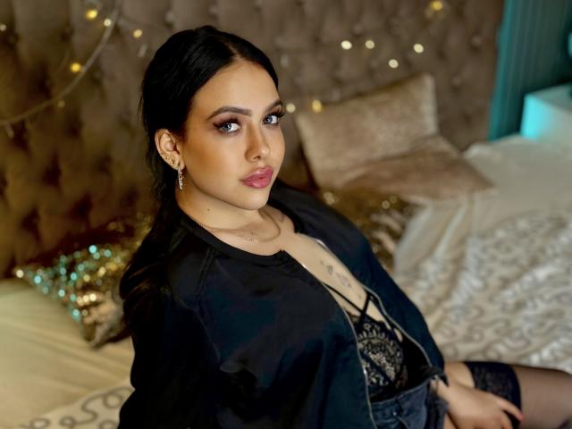 MilanaJuice, con cabello negro, vestida con una chaqueta negra, un sujetador de encaje y aretes, sentada en una cama, mirando a la cámara con una ligera sonrisa, en un dormitorio..