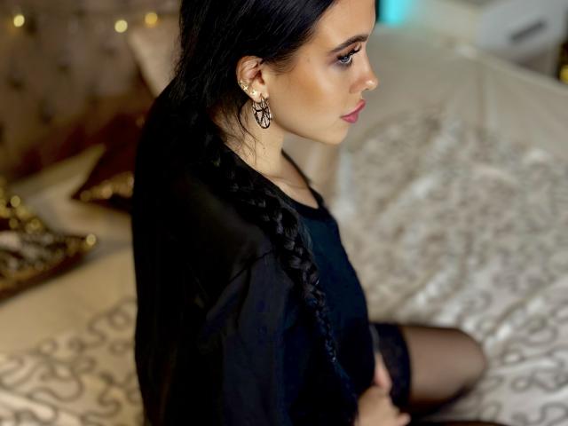 Milana, con cabello largo y negro trenzado, vestida con una camisa negra, sentada en una cama, mostrando sus medias negras que le llegan al muslo, y con aretes elaborados, mirando hacia un lado..