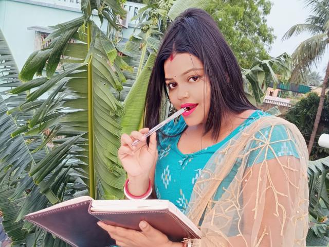 Surovi with dark hair, blue dress, and red bindi, licking a pencil, holding a notebook, green foliage in background, sheer gold patterned shawl, red bangle, looking cute.