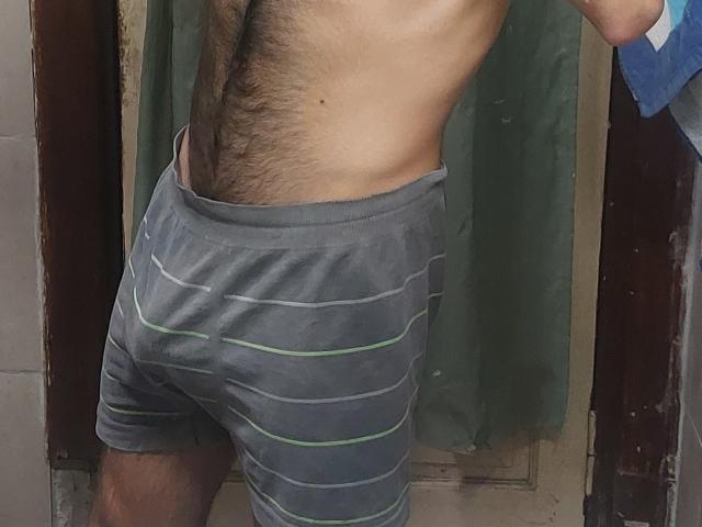 LapijArgie69's bare, hairy, tan-skinned torso in gray striped shorts, showing a slight ass curve and a hand in his pocket, standing near a green curtain.