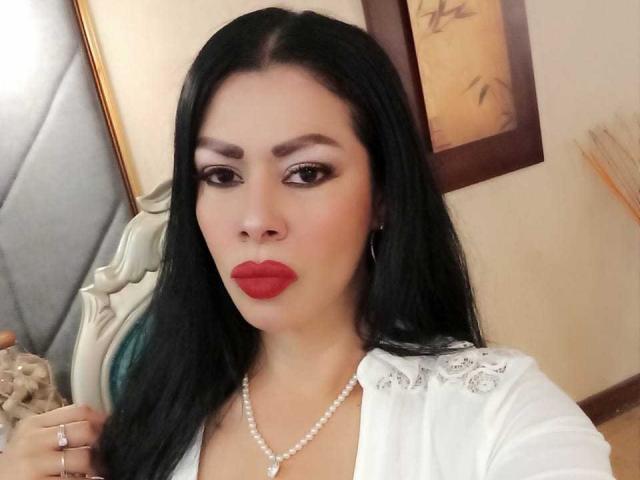 SofiMontes a de longs cheveux noirs, du rouge à lèvres et un collier de perles. Elle porte une chemise blanche avec de la dentelle, et ses yeux marrons sont fixés sur l'appareil photo..