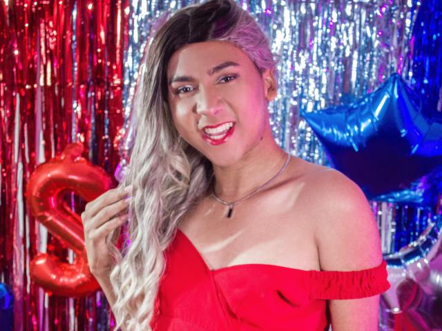 ZoeHotMoretti with long, wavy blonde hair, red off-shoulder dress, smiling, holding hair, wearing necklace, shiny red and silver background, blue star balloons, looking cute.