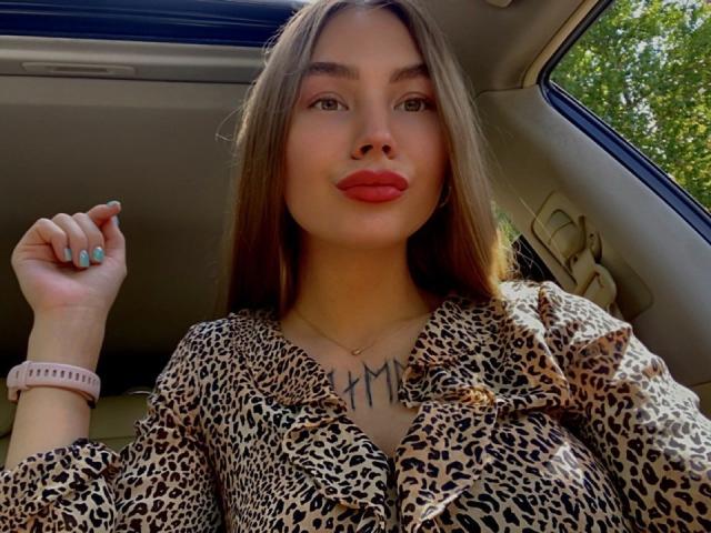 LexiLee with long brown hair, red lipstick, leopard print shirt, tattoo on chest, pink wristband, holding hair, sitting in car, sunny day, green tree outside window.