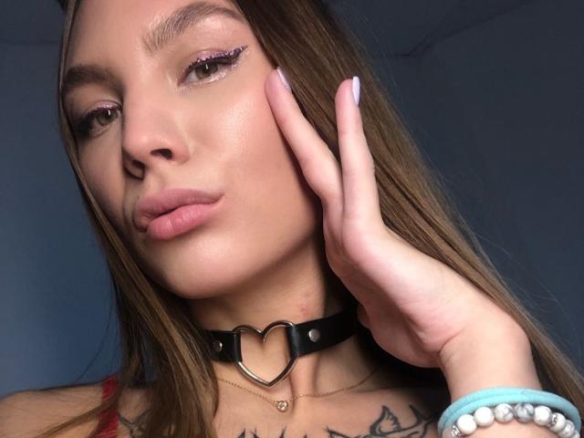 LexiLee has light brown skin, long brown hair, pink eyeliner, pink lips, wearing a black heart choker, touching her cheek with pale pink nails, and a turquoise bracelet.