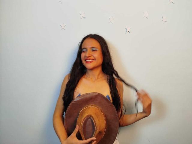 CrisyMarie with long dark hair, smiling, holding a brown cowboy hat, touching her hair, wearing a blue bra, showing her tits, against a plain light wall.