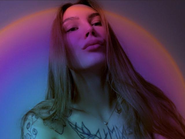 LexiLee with long brown hair, tattoos on her chest, and a confident expression, lit with purple and pink neon lights. Her boobs are partially visible, topless.