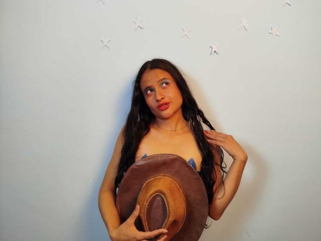 CrisyMarie with long black hair, holding a brown cowboy hat, covering her small tits with her hands, wearing blue straps, red lipstick, light skin, white wall with star stickers.
