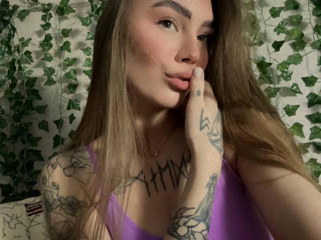 LexiLee with long brown hair, pink lips, and tattoos on her arm and chest, wearing a purple top, touching her cheek with a finger, looking at the camera.