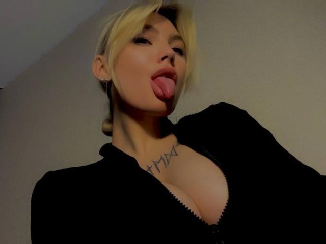 LexiLee has blonde hair, her tongue out, and is wearing a black zip-up top showing her tits. She has a triangle tattoo on her chest.