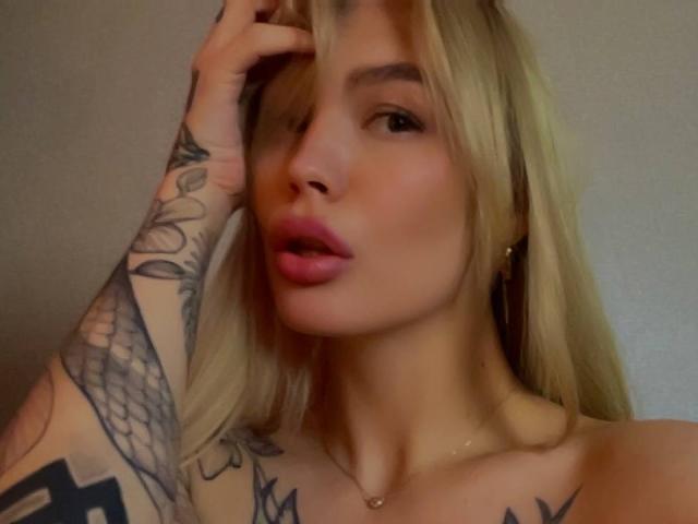 LexiLee has blonde hair, pink lips, and tattoos on her arm. She's touching her head with a hand, showing off her tattoos and necklace. Boobs visible, topless.