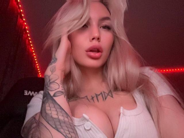 LexiLee with platinum blonde hair, full lips, and tattoos, wearing a low-cut white top, showing big tits, holding her head, red LED strip lights in the background.