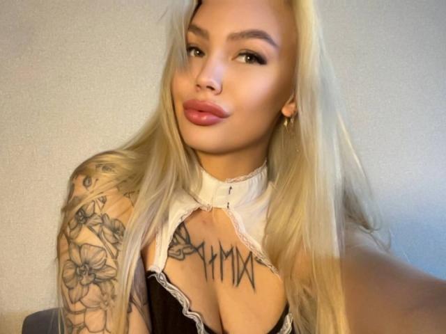 LexiLee met lang blond haar, tatoeages op haar arm en borst, gekleed in een zwart-wit top, met een glimlach en roze lippen, en gouden oorbellen. Haar borsten zijn zichtbaar op de foto..