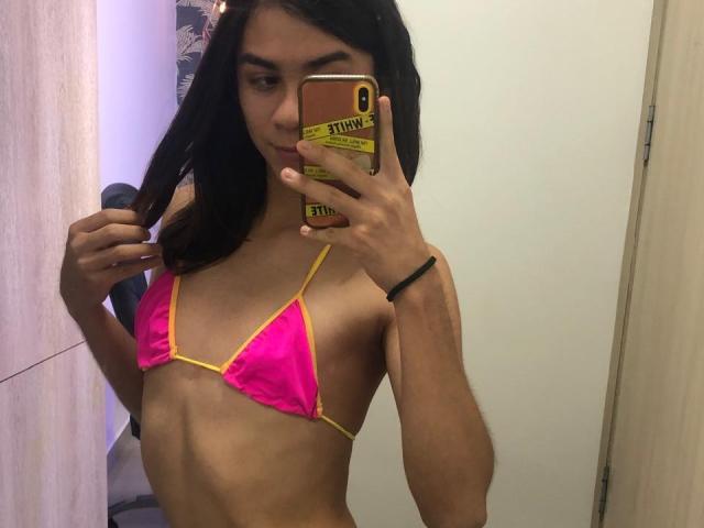 TamaraGarcia in a pink and yellow bikini, showing a slim, flat-chested, androgynous upper body with dark hair, holding a phone in a mirror selfie.