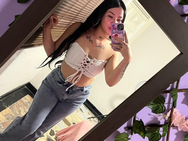 EmmieX with long black hair, white strapless top showing small tits, lace-up front, blue jeans, tattoos, holding phone, mirror selfie, pink lipstick, purple wall, wooden floor.