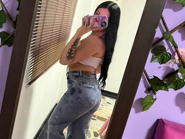 EmmieX with long black hair, wearing a pink tube top and high-waisted blue jeans, showing her ass in a mirror selfie, holding a pink phone with a sticker.